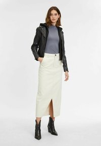 Schwarze Leder-Bikerjacke, graues eng anliegendes Oberteil, cremefarbener Midirock mit vorderem Schlitz und schwarze Stiefel mit Absatz und strukturierter Oberfläche.