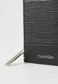 Portefeuille en cuir noir avec une finition texturée, quincaillerie en argent et logo "Calvin Klein" gaufré sur un accent noir lisse.