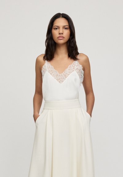 Camisole en satin blanc avec un décolleté en dentelle, associé à une jupe ivoire taille haute avec des plis et des poches latérales.