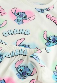 Valkoinen kangas, jossa on värikkäitä Stitch-hahmon printtejä ja sana "OHANA" mustalla. Materiaalilla on pehmeä, teksturoidun näköinen pinta.