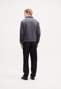 Young man stands facing away, wearing a dark gray jacket, black pants, and black shoes against a plain white background.