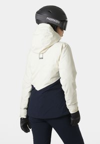 Ski jacket featuring a white upper portion with a navy lower section, a hood, and black gloves. The design includes a prominent logo on the back.