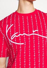 Röd bomulls-t-shirt med vita ankarmönster och en framträdande vit broderad logotyp. Kort ärm och rundad halsringning.