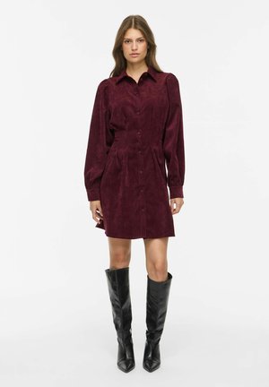 Femme portant une robe bordeaux à manches longues boutonnée avec un col et des bottes en cuir noires montant jusqu'aux genoux, debout sur un fond blanc.