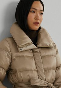 Beige pufferjack met een hoge kraag, ritssluiting en ceintuur in de taille. De stof heeft een gladde, glanzende textuur met horizontale quilt.