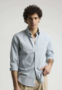 Camisa de botones de color azul claro con un patrón de rayas. Presenta un cuello, mangas arremangadas y una etiqueta en el dobladillo. Textura de tela suave.