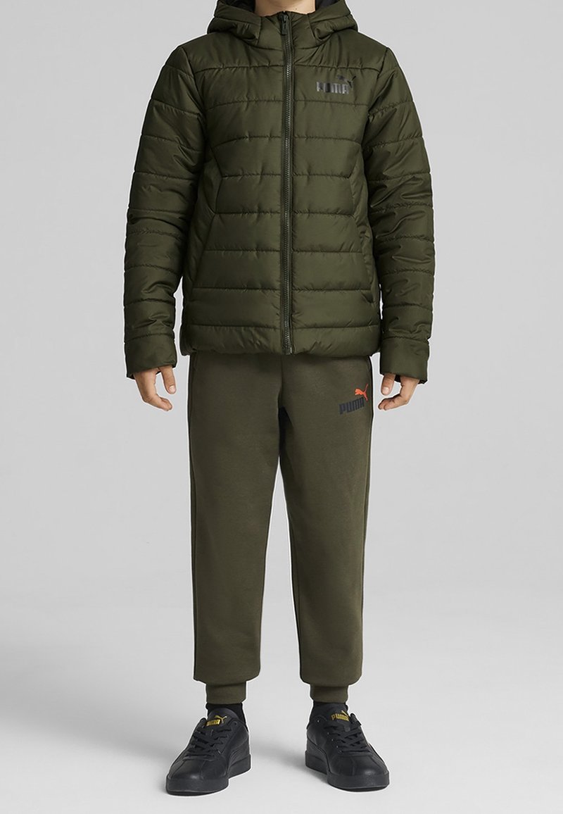 Enfant portant une doudoune kaki avec capuche, un pantalon de survêtement Puma assorti et des baskets noires Puma, debout devant un fond uni.
