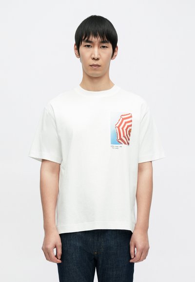 T-shirt en coton blanc avec une illustration carrée d'un parapluie rayé rouge et blanc et le texte "Good things and tan lines" sur la poche.