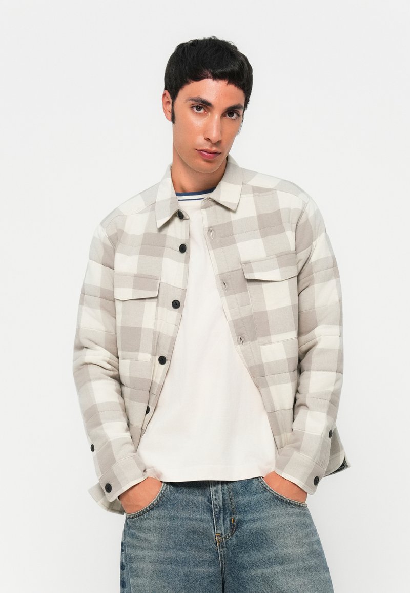 Junger Mann, der eine beiger-weiße karierte Hemdjacke über einem weißen T-Shirt trägt, mit den Händen in den Taschen seiner blauen Jeans.