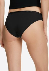 Kobieta w czarnych majtkach w stylu hipster, z elastycznym paskiem z logo, pokazana od tylnej strony na wysokości talii do połowy uda.