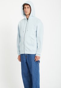 Veste à capuche zippée bleu clair avec une capuche, poches avant et quincaillerie en métal argenté. Associée à un jean en denim bleu, ample. Texture du tissu lisse.
