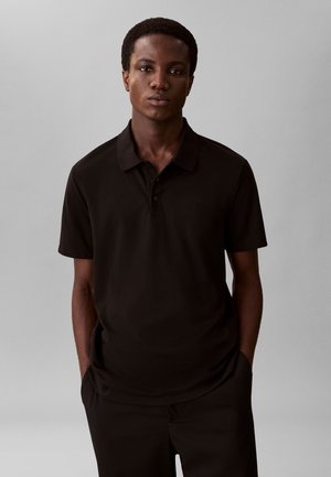 Bărbat tânăr cu păr scurt și creț, purtând un tricou polo negru și pantaloni, stând cu mâinile în buzunare, pe un fundal gri simplu.