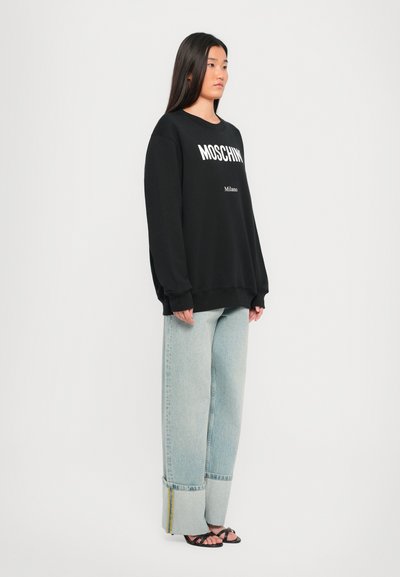 Sudadera negra de gran tamaño con el logotipo blanco "MOSCHINO Milano", combinada con unos vaqueros de pierna ancha de color azul claro con dobladillo enrollado y un acento amarillo.