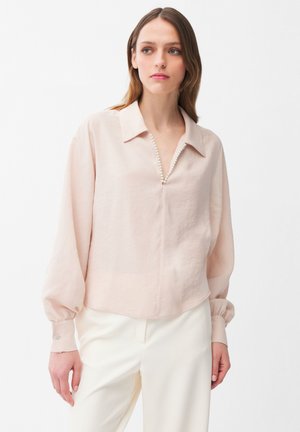 Kobieta w jasnoróżowej bluzce z perłową lamówką przy dekolcie w serek, długimi bufkami na rękawach oraz białych spodniach na jednolitym tle.