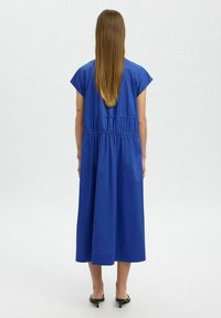 Blaues, knielanges Kleid mit kurzen Ärmeln, tailliertem Schnitt und glatter Textur. Getragen mit schwarzen, hohen Sandalen, von hinten betrachtet.