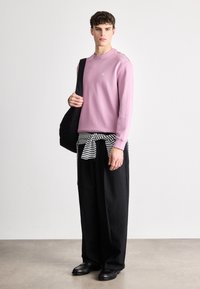 Sudadera de manga larga en un suave color rosa claro con un pequeño logo, combinada con un bolso negro y pantalones holgados negros, sobre un envoltura de rayas en blanco y negro.