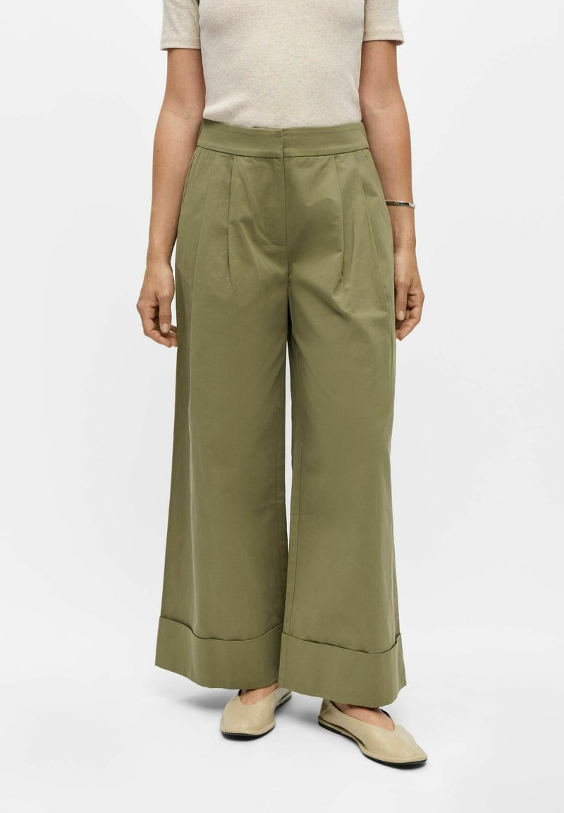 Personne portant un pantalon large taille haute vert olive, un haut beige à manches courtes, des ballerines beiges et un bracelet argenté au poignet droit.