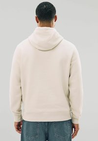 Sudadera con capucha beige hecha de una tela suave, con un bolsillo canguro y puños acanalados, combinada con unos vaqueros de color azul claro.