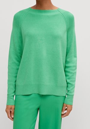 Person trägt einen einfarbigen grünen Pullover mit langen Ärmeln zusammen mit passenden grünen Hosen vor einem einfarbigen hellen Hintergrund.