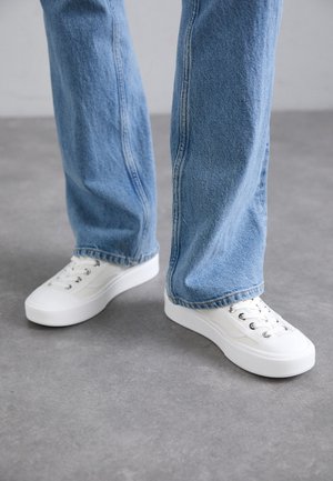 Persona che indossa jeans azzurri a gamba dritta e sneakers bianche basse con lacci, in piedi su pavimento grigio.