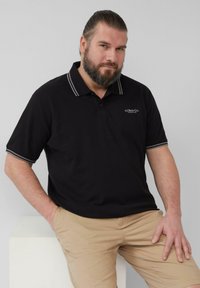 Zwarte polo met een grijsgestreepte kraag en manchetten, met een klein geborduurd logo. Draag het met lichte beige broek tegen een neutrale achtergrond.
