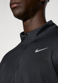 Svart Nike zip-up jacka med hög krage och en silverfärgad logotyp på vänster bröst. Tyget har en slät, texturerad yta.