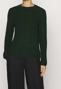 Maglione lavorato a maglia verde scuro con un motivo testurizzato, scollatura rotonda e polsini a costine. Abbinato a pantaloni neri a gamba larga.