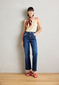 Top a coste beige, jeans aderenti blu scuro a vita alta con cuciture dorate, e sandali con tacco alto rossi con lacci. Modella in piedi con le mani in tasca.