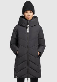 Zwarte pufferjas met capuchon, voorzien van een rits aan de voorkant, chevronpatroon en een logotag. Geribde zwarte beanie op het hoofd van het model.