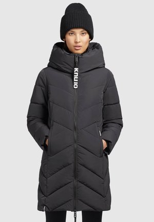 Piumino nero con cappuccio, dotato di zip frontale, motivo a chevron e etichetta con logo. Berretto nero a costine sulla testa del modello.