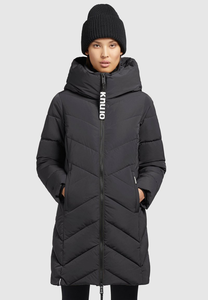 Zwarte pufferjas met capuchon, voorzien van een rits aan de voorkant, chevronpatroon en een logotag. Geribde zwarte beanie op het hoofd van het model.