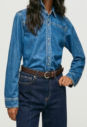 Person iført en blå denimskjorte med knapper, stoppet ned i mørkeblå jeans med et brunt læderbælte, hånd hvilende på hoften.
