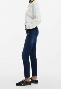 Tummansiniset slim-fit farkut, joiden taskuissa on kukka-embroidery, yhdistettynä kermaiseen palmikkoneuleeseen ja mustiin tennareihin, joissa on valkoisia yksityiskohtia.