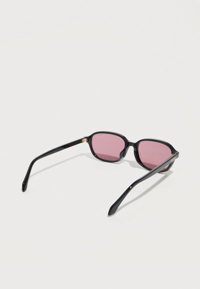 Μαύρα ορθογώνια γυαλιά ηλίου με ροζ φακούς τοποθετημένα σε λευκή επιφάνεια, κεκλιμένα ώστε να φαίνονται και τα δύο μπράτσα και οι φακοί.