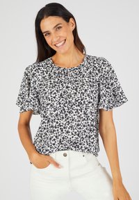 Bloemrijke top met korte mouwen in het wit met een zwart patroon. Heeft een geplooide halslijn en een ontspannen pasvorm. G gedragen met lichtgekleurde jeans.