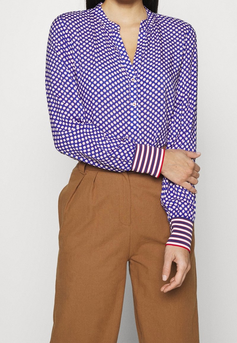 Blusa de manga larga morada con lunares blancos, cierre de botón y puños a rayas rojas. Combinada con pantalones de cintura alta marrones.