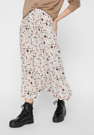 Femme portant une jupe midi blanche à imprimé floral avec un ourlet volanté, un haut beige à manches longues et des bottines noires à plateforme lacées, debout devant un fond uni.