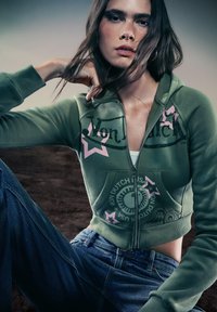 Groene zip-up hoodie met roze ster graphics en logo print. Zachte, katoenen blend materiaal met geribbelde manchetten en zoom. Gecombineerd met blauwe spijkerbroek.