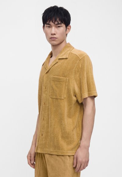 Jeune homme aux cheveux courts noirs porte une chemise moutarde à manches courtes boutonnée et un pantalon assorti sur un fond blanc uni.