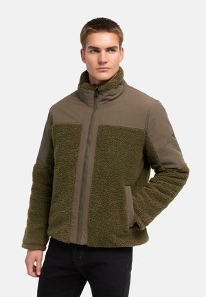 Giovane uomo che indossa una giacca in pile verde con zip frontale e colletto alto, guarda leggermente di lato, mani nelle tasche.