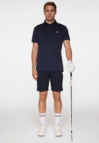 Mörkblå pikétröja med krage, tillsammans med matchande mörkblå shorts. Vit golfhandske på vänster hand, håller i en golfklubba. Vita sneakers och randiga strumpor.