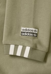 Light olive green Adidas sweatshirt cuff with white three-stripe detail and dual black and white Adidas logo patches.