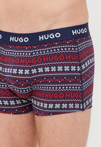 Marineblaue Boxershorts mit rotem und weißem festlichem Muster, das Rentiere, Schneeflocken und den wiederholten "HUGO"-Schriftzug auf dem Bund zeigt.