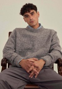 Pull en laine gris avec un col montant, et une chemise bleu clair à col en dessous. Modèle assis, mains jointes, portant un pantalon gris clair.