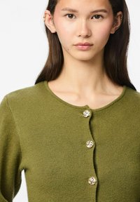Olive zaļš kardigans, izgatavots no mīkstas auduma, ar apaļu izgriezumu un dekoratīvām zelta krāsas ziedu pogām priekšpusē.