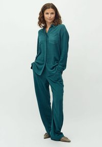 Completo due pezzi color teal composto da una camicia con bottoni e due tasche sul petto, abbinata a pantaloni larghi. Tessuto morbido con una consistenza liscia.