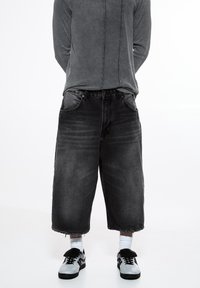 Culottes en denim noir à coupe décontractée, avec des ourlets effilochés et un design classique à cinq poches, associées à des baskets noires et des chaussettes blanches.