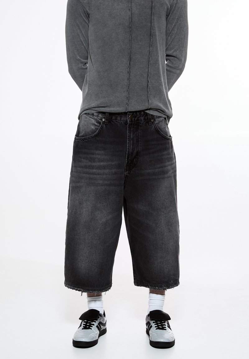 Culottes en denim noir à coupe décontractée, avec des ourlets effilochés et un design classique à cinq poches, associées à des baskets noires et des chaussettes blanches.