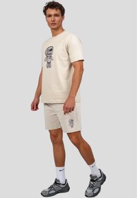 Camiseta de algodón beige con un estampado gráfico que presenta a un astronauta caricaturesco. Combinada con pantalones cortos beige a juego, ambos ofrecen un ajuste relajado.