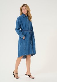 Denimmekko, jossa on kaulus, pitkät hihat ja sidottava vyötärö. Ominaisuuksina sininen väri, taskut ja polven alapuolelle ulottuva, kaareva helma.
